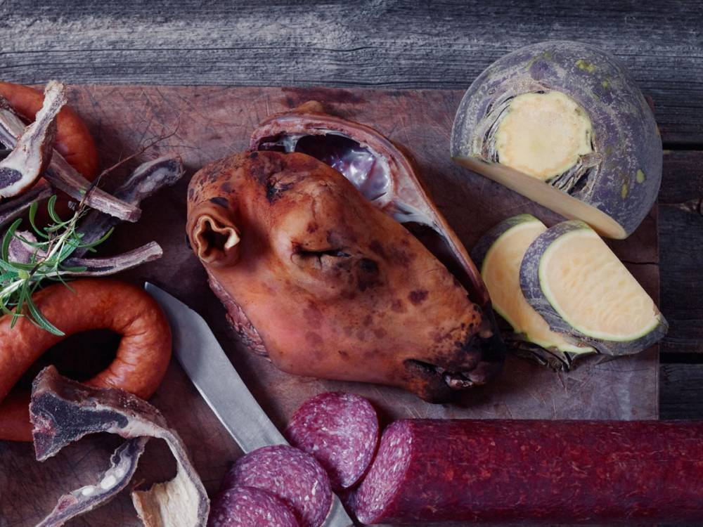 Smalahove med tilbehør som pølse, ost og smør på et trebenk.