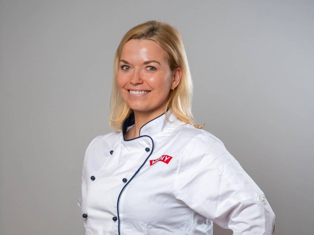 En kvinne i hvit kokkekjole med logo står med armene i kors, mot en grå bakgrunn.