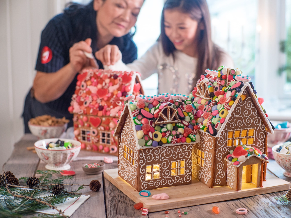To personer lager et pepperkakehus med fargerike dekorasjoner og godteri på et bord.