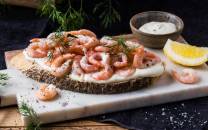 Rekesmorbrod med reker, dill, sitron og en liten skål med hvit saus på en skjærebrett.