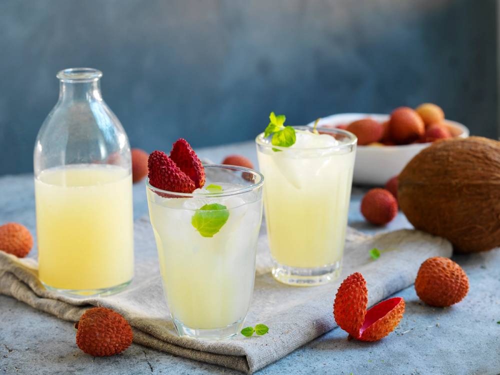 To glass med lyse gul limonade med litchi-frukter og mynte, omgitt av flere litchi-frukter og en flaske limonade på et kjøkkenbord.