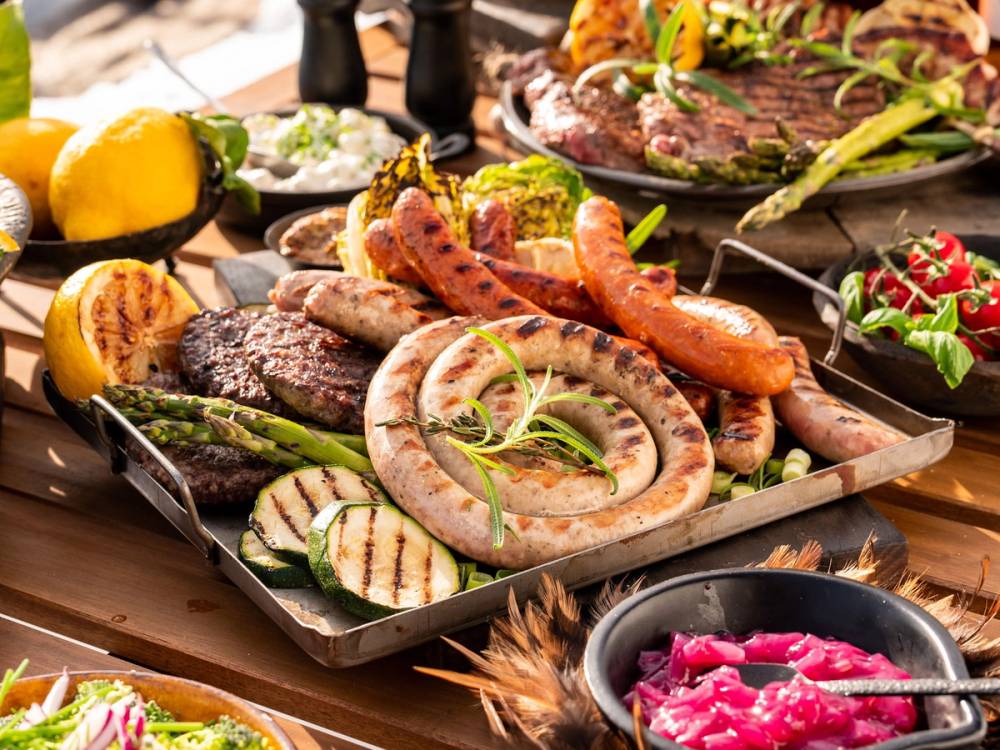 Et utvalg av grillet kjøtt, pølser og grønnsaker på et serveringsfat, med sitronbåter og grønne urter som pynt.