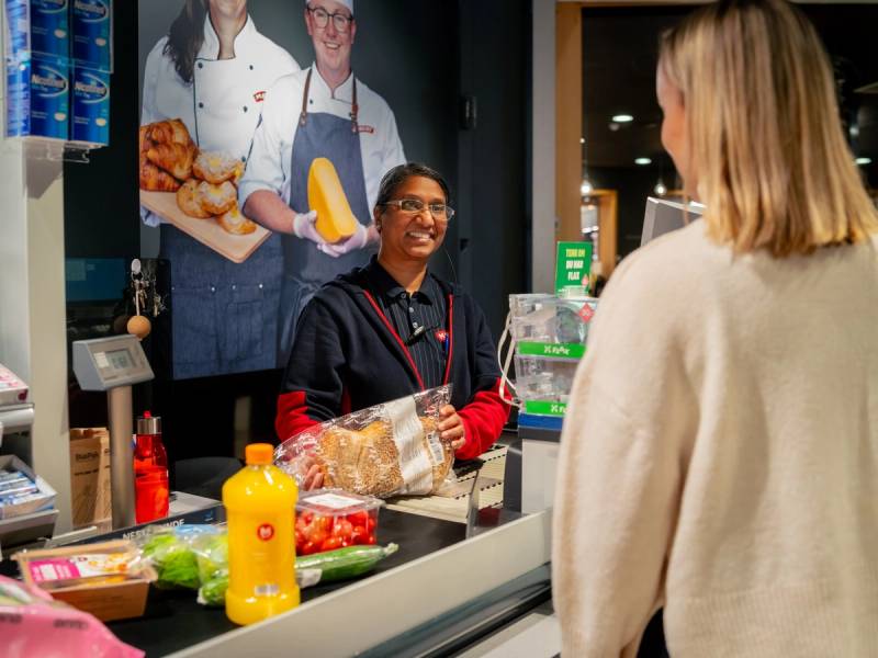 Kunde i kasseområdet på MENY Løren med en ansatt som pakker varer, mens en annen kunde venter foran i køen.