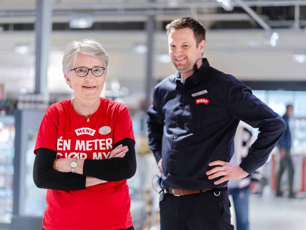 To personer står sammen i et innendørs miljø, en av dem har på seg en rød t-skjorte med tekst, den andre en mørk jakke, og de har begge armene i kors.