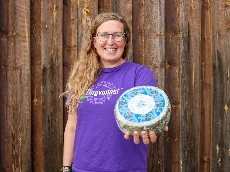 En person i lilla t-skjorte holder en stor blå og hvit ostekule foran en trevegg.