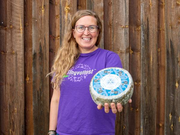 En person i lilla t-skjorte holder en stor blå og hvit ostekule foran en trevegg.