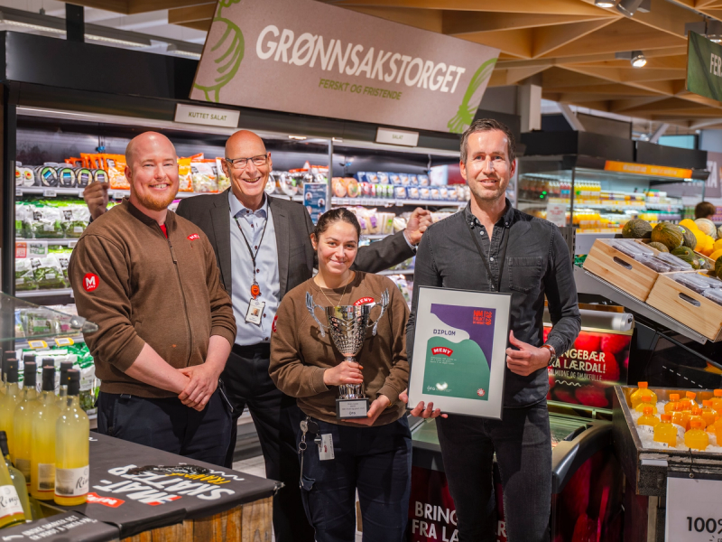 En gruppe mennesker står i en butikk, og en av dem holder et trofé, mens en annen viser frem et bilde på en plakat, med frukt og grønnsaker i bakgrunnen.
