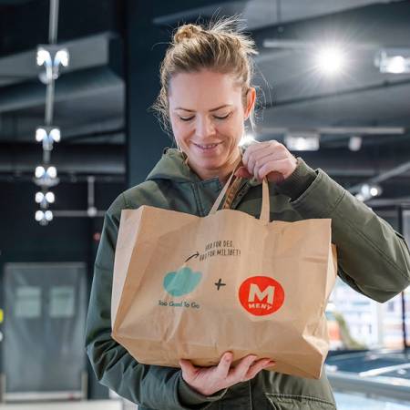 En person holder en papirpose med mat fra Too Good To Go i en butikk eller et kjøpesenter.