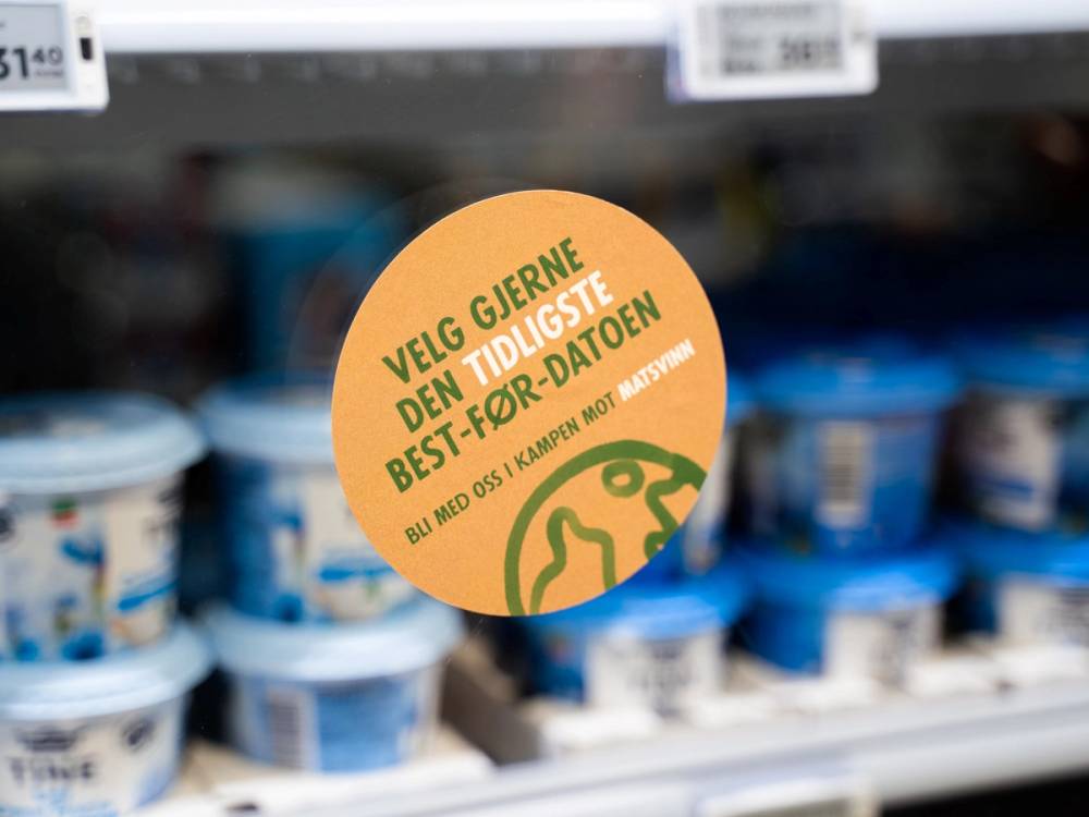 En plakat med teksten 'Vei gjerne den tildigste best for deg' plassert blant kjølevarer i en butikk, sannsynligvis meieriprodukter.