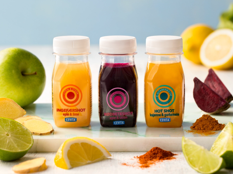 Tre små flasker med ulike farger på juice eller smoothie, omgitt av frukt og krydder, på et lyst bord.