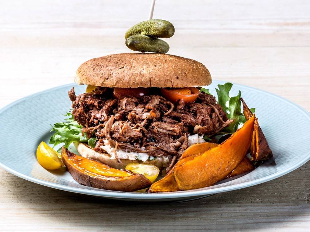 En burger med pulled pork, serveres med ovnsbakte gresskarbiter og salat på en blå tallerken.