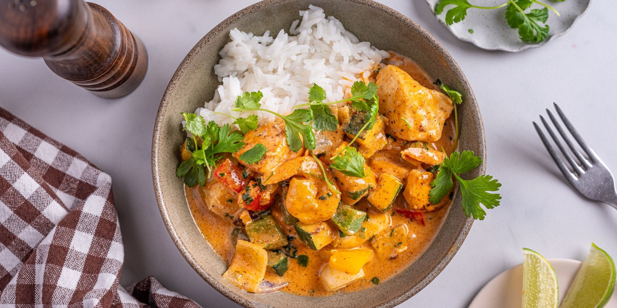 Fisk wok med rød curry servert med ris i en skål, pyntet med frisk koriander, på et bord med bestikk og en limebåt.