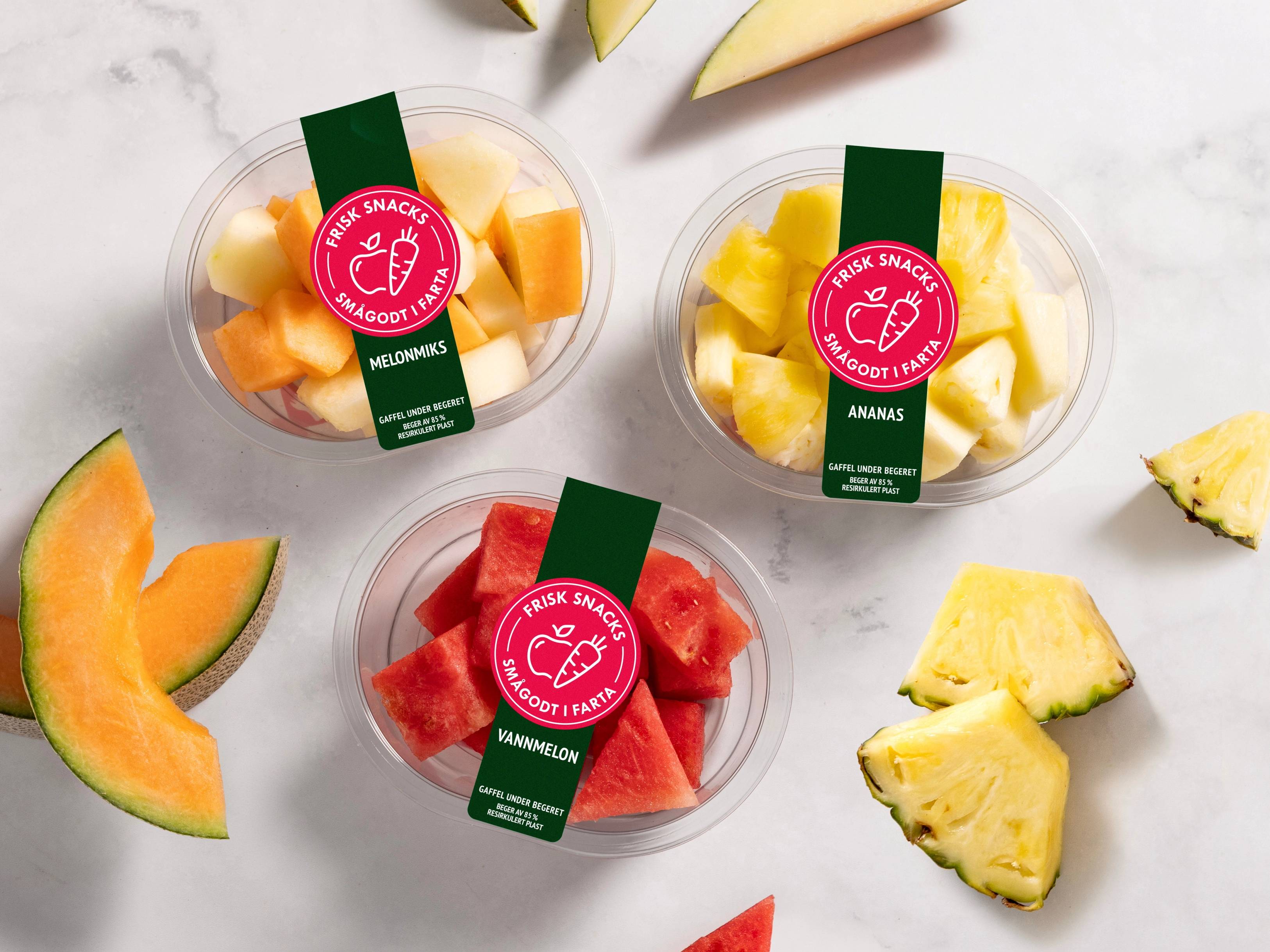 Fargede biter av frukt i små plastkopper med etiketter, omgitt av fruktstykker på en hvit bakgrunn.