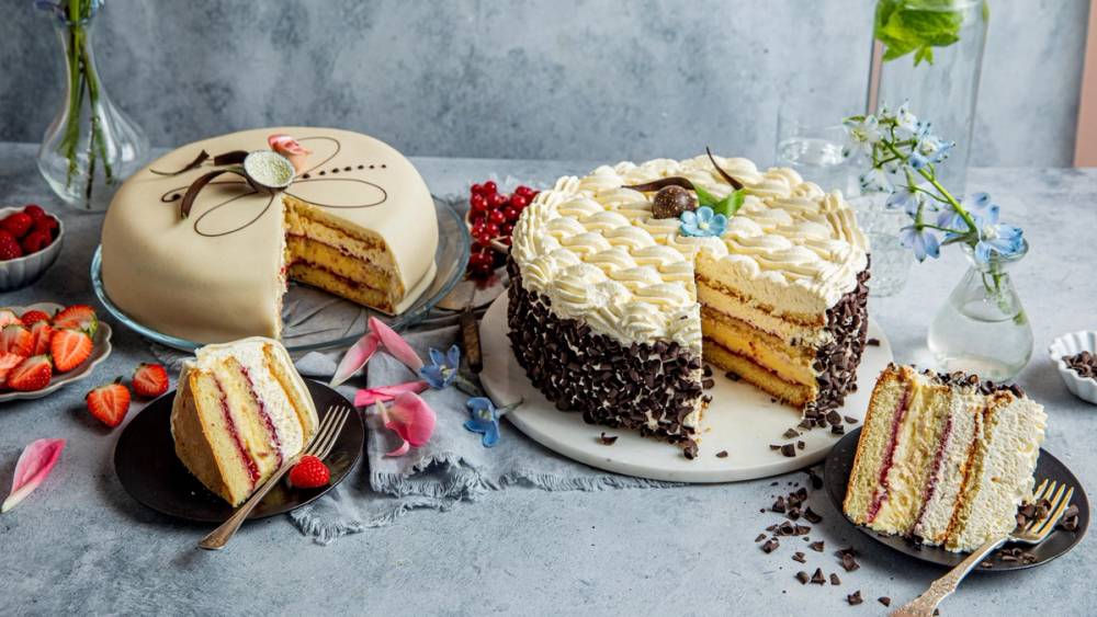 To kaker, en med hvit krem og sjokoladekrøller, og en annen med gul krem og rosa dekor, omgitt av jordbær og blomster på et bord.