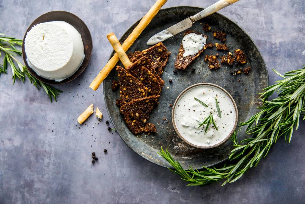 Ricottaost i en liten skål med urter, omgitt av brød og frisk rosmarin på en steinplate