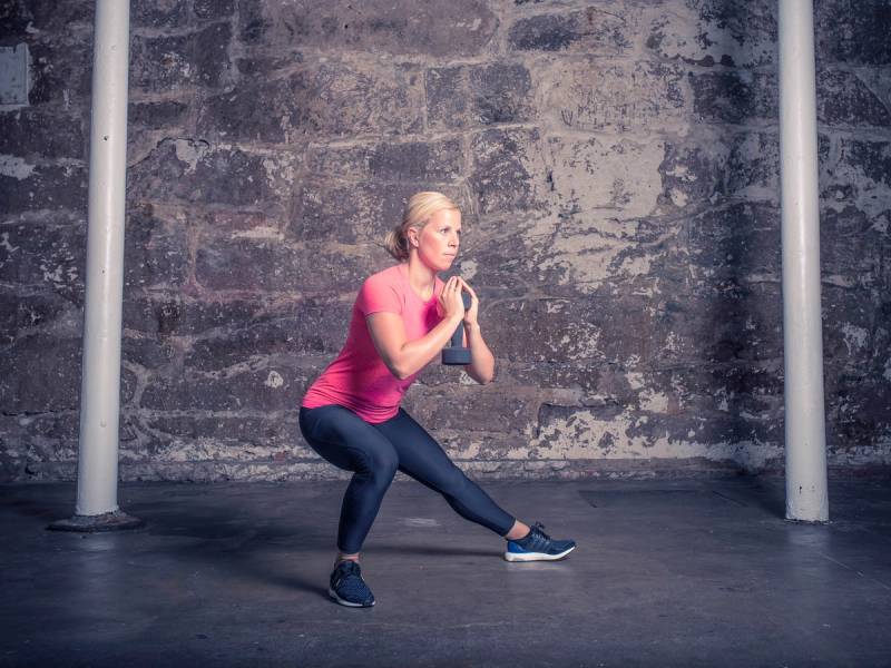En person i rosa treningtøy gjør en sideutfallsøvelse mot en murvegg, med armer i bønneposisjon foran brystet.