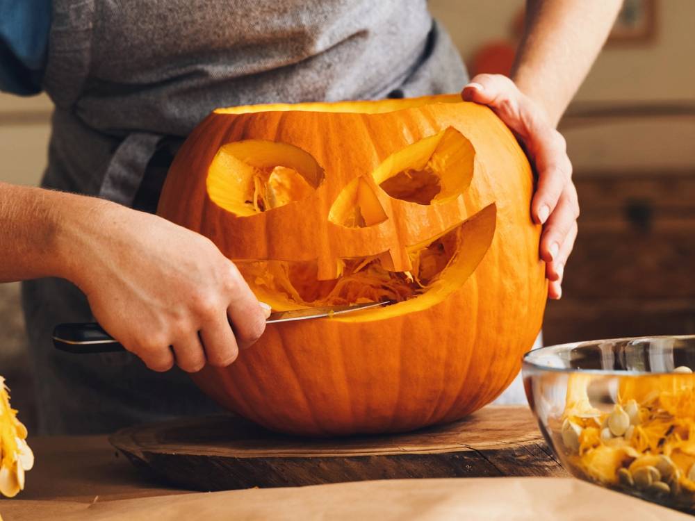 Person som skjærer ut ansikt og øyne i en gresskar til Halloween, med en bolle med utskåret gresskarkjøtt i nærheten.