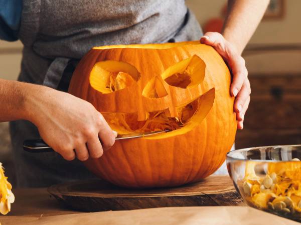 Person som skjærer ut ansikt og øyne i en gresskar til Halloween, med en bolle med utskåret gresskarkjøtt i nærheten.
