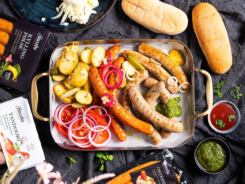 Et utvalg av pølser, potetsalat, grønnsaker og tilbehør på et serveringsfat, med brød og saus rundt.