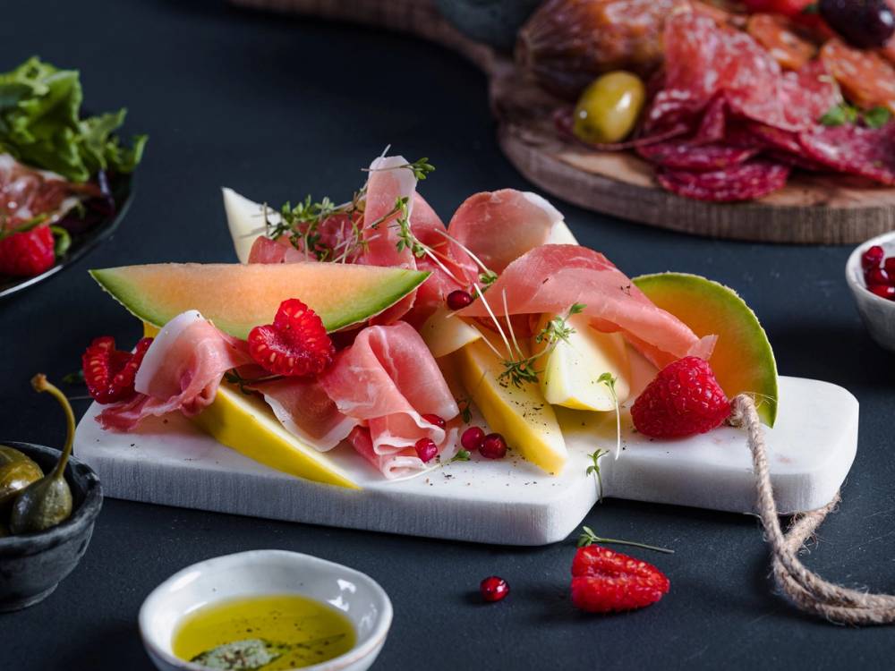 Spekeskinke og melon på et serveringsfat med friske bær og urter, omgitt av små skåler med saus og olivenolje.