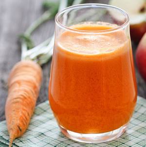 Et glass med oransje juice eller smoothie med en gulrot i bakgrunnen, plassert på et kjøkkenbord.