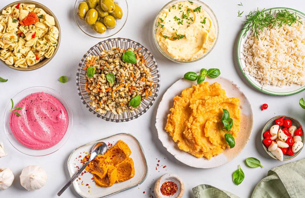 Et utvalg av ulike sauser og tilbehør på tallerkener og skåler, inkludert hummus, ris, grønnsaker og dressing, på et bord.