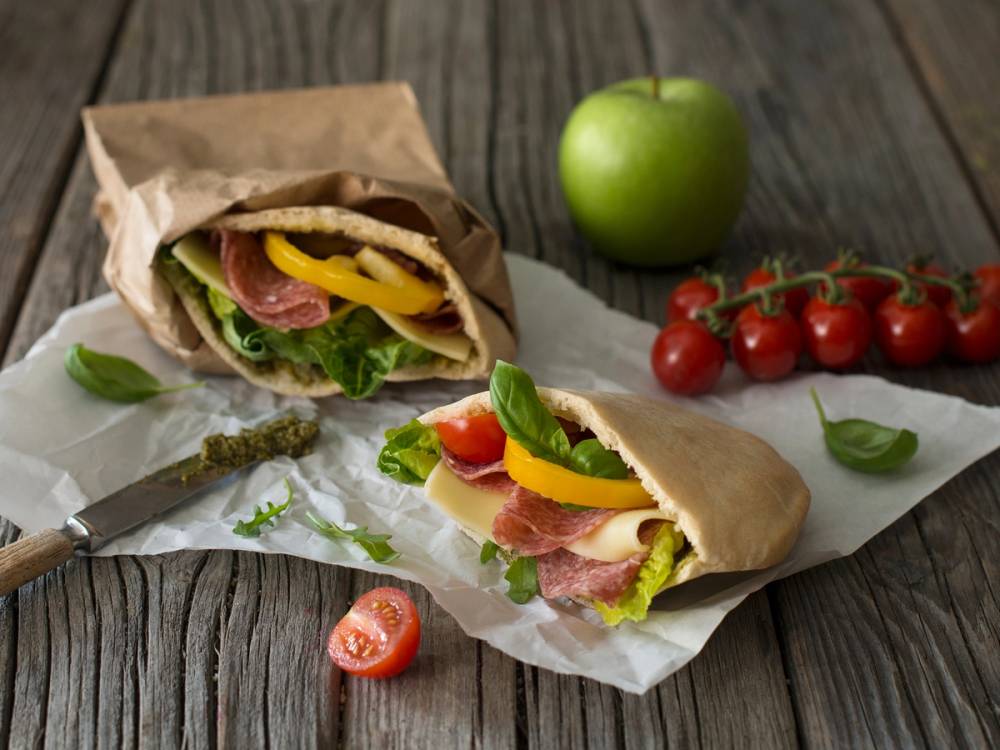 Pita fylt med salami, gulost og pesto, med en grønn eple og cherrytomater ved siden av på et trebord.