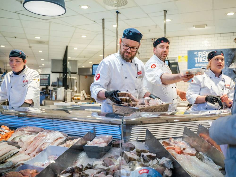 Kjøkkensjefer i hvite uniformer og blå hodeplagg jobber bak en fiskedisken i en matbutikk.