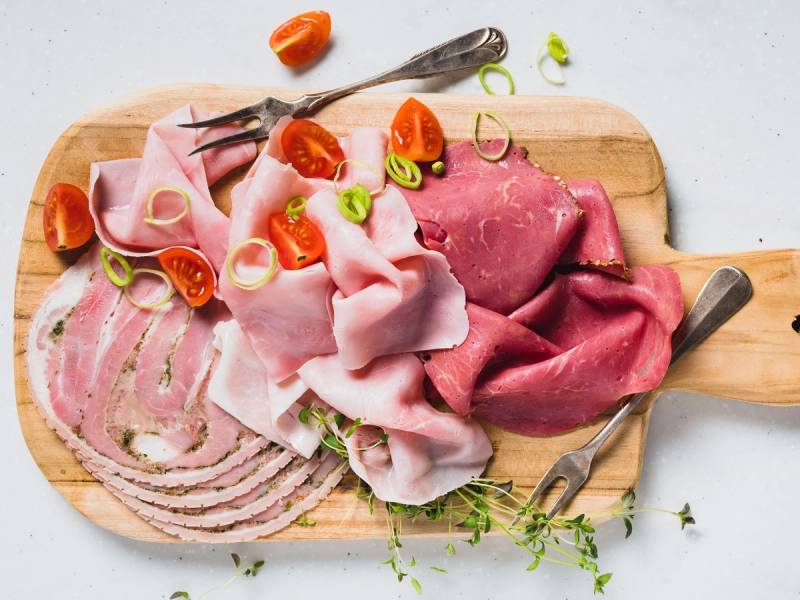 Ulike typer av kjøtt og skinke på en skjærebrett med grønnsaker og krydder, klar for tilberedning.