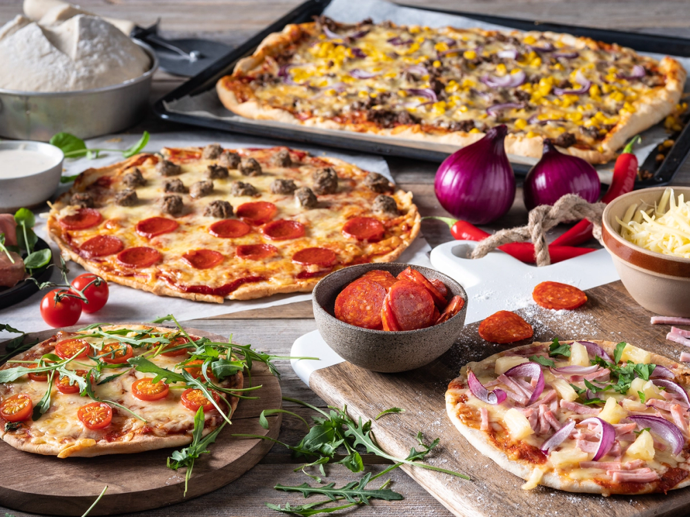 Ulike pizzaer med ulike pålegg, inkludert pepperoni, grønnsaker og ost, på et bord med ingredienser og tilbehør.