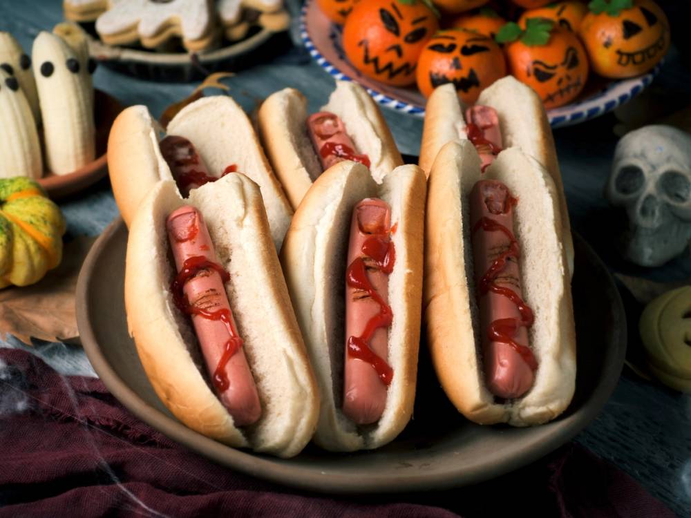 Tre hot dogs med ketchup i hvite brød, omgitt av halloween-dekorasjoner som små gresskar og skjeletter.