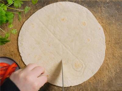 En person skjærer i en rund tortilla på et trebenk, med litt grønt og en liten tallerken med paprika i nærheten.