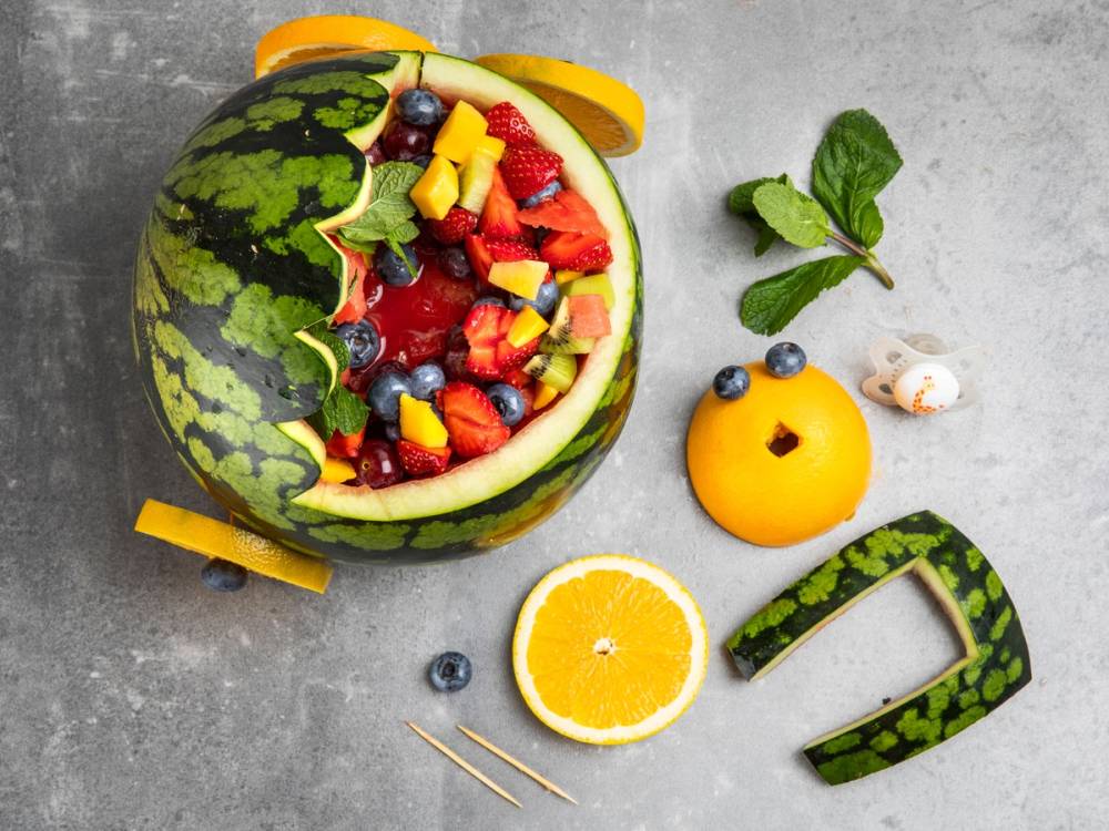 En vannmelon fylt med ulike frukter som jordbær, blåbær, mango og melon, omgitt av en sitronhalvdel, en liten gul vannmelon, og noen mynteblader på en grå overflate.