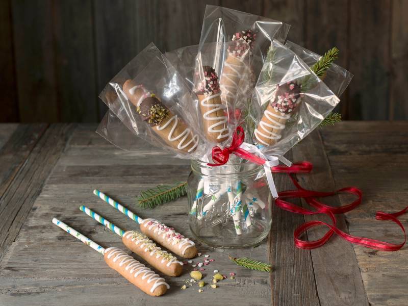 Kransekakestenger dekorert med sjokolade og strøssel, pakket inn i cellofan og bundet med rødt bånd, plassert i en glasskrukke på et bord.