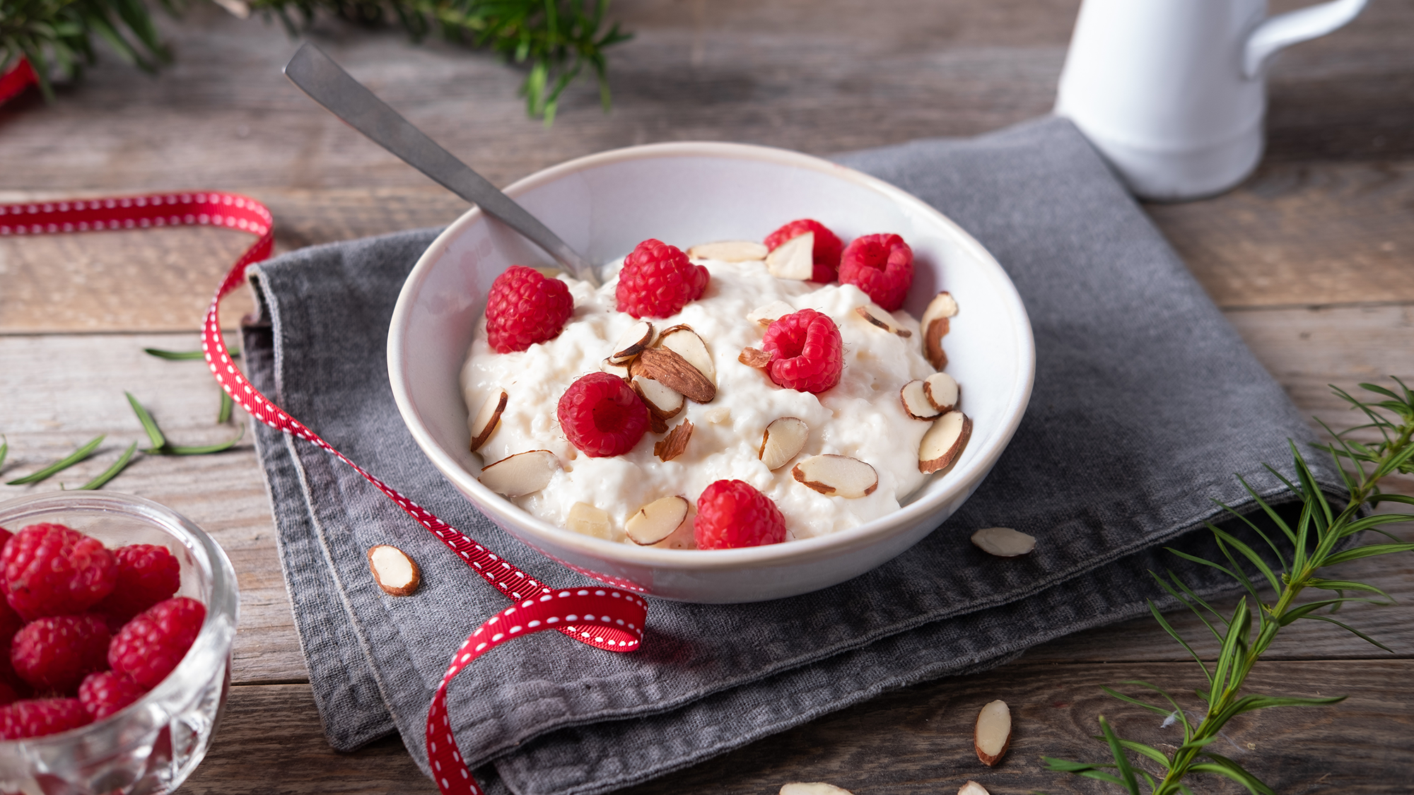 Risgrøt med ripsbær og skivede mandler i en hvit bolle på et bord med en skje oppi, omgitt av grønne planter og en liten skål med flere ripsbær.