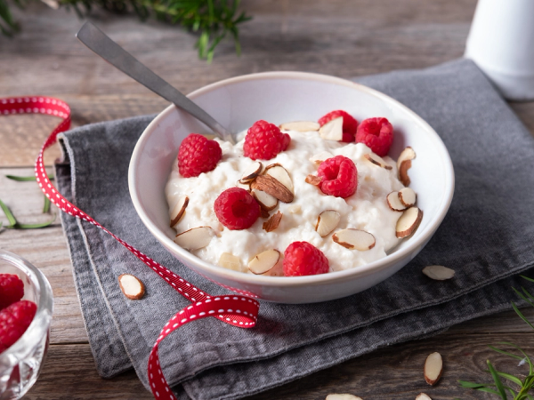 Risgrøt med ripsbær og skivede mandler i en hvit bolle på et bord med en skje oppi, omgitt av grønne planter og en liten skål med flere ripsbær.