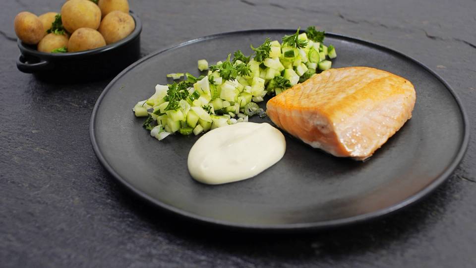 Pannestekt laks med en klatt med hvit saus og en side av hakkede grønnsaker på en svart tallerken.
