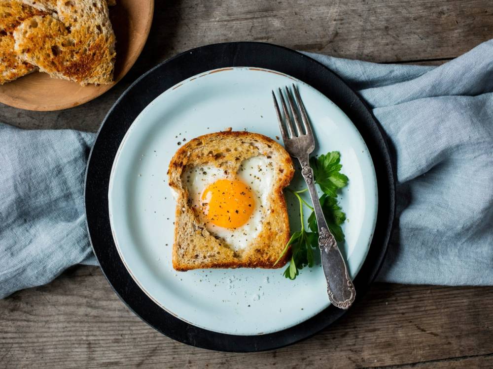 Stekt egg på en skive brød med en liten grønn urt ved siden av på en hvit tallerken, med en gaffel og en blå duk ved siden av.