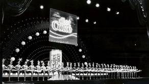 En gruppe ballettdansere i tutu står i en rekke på scenen under en forestilling med en stor Coca-Cola-logo i bakgrunnen.