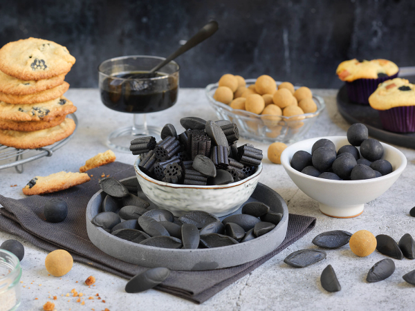 Et utvalg av lakrisgodteri, inkludert svarte lakrisskiver, lakrisbåter og lakrisskudd, på et bord med en liten skål med lakrissirup og noen kjeks i bakgrunnen.