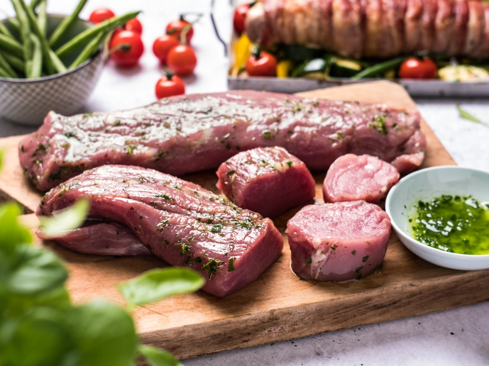 Rå svinefilet og indrefilet på et skjærebrett med marinade og urter, omgitt av grønnsaker og tomater.