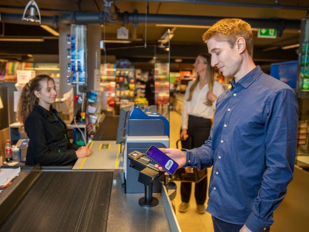 En mann i blå skjorte betaler med mobiltelefon i kasse i butikk, mens en ansatt hjelper til i bakgrunnen.