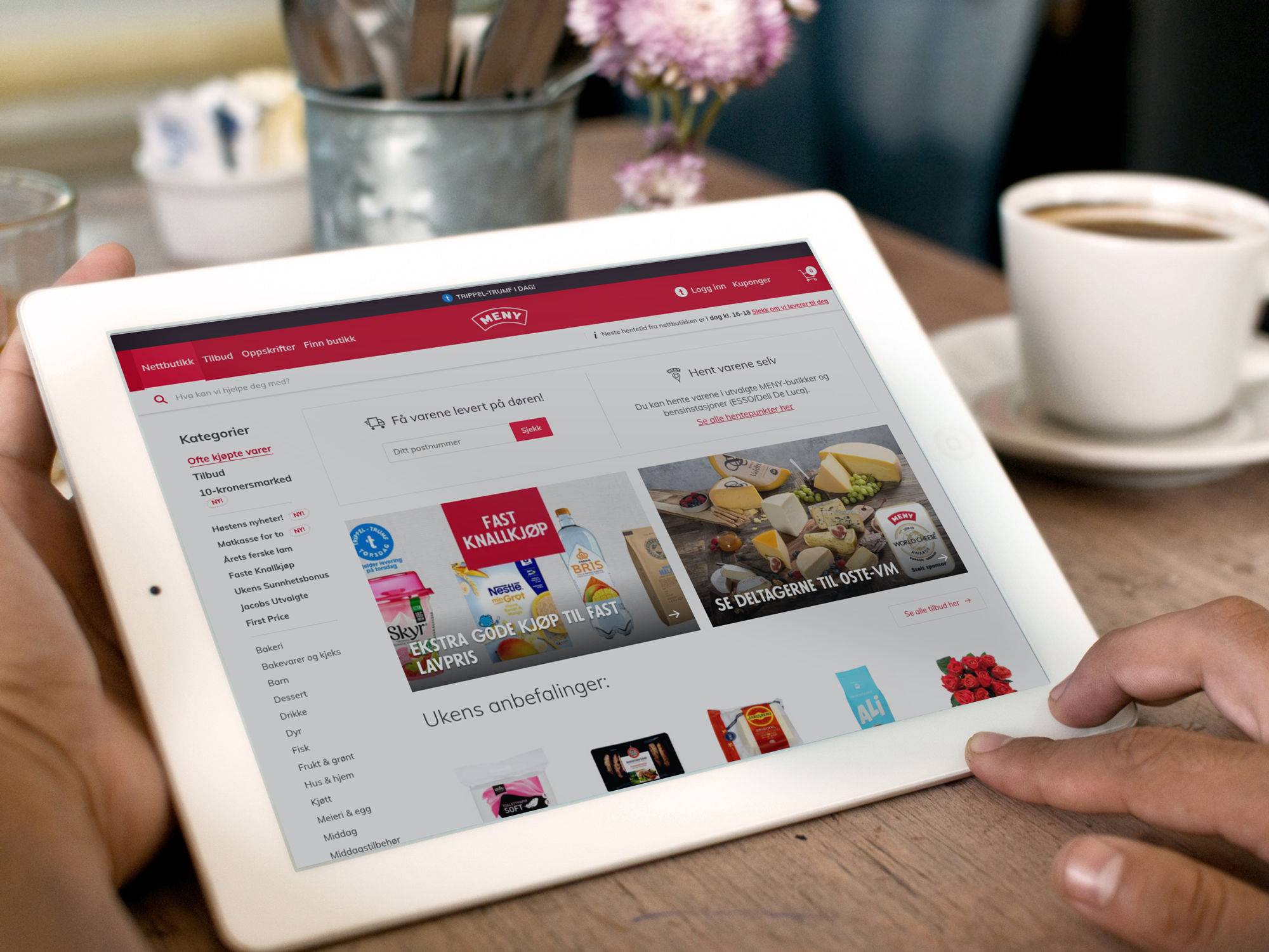 En person bruker en nettbrett med en nettside åpen, mens det er en kopp kaffe og en blomsterbukett i bakgrunnen.
