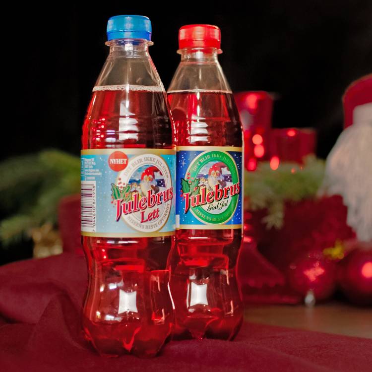 To flasker med Ringnes julebrus, en med blått lokk og en med rødt lokk, står på et bord med julepynt i bakgrunnen.