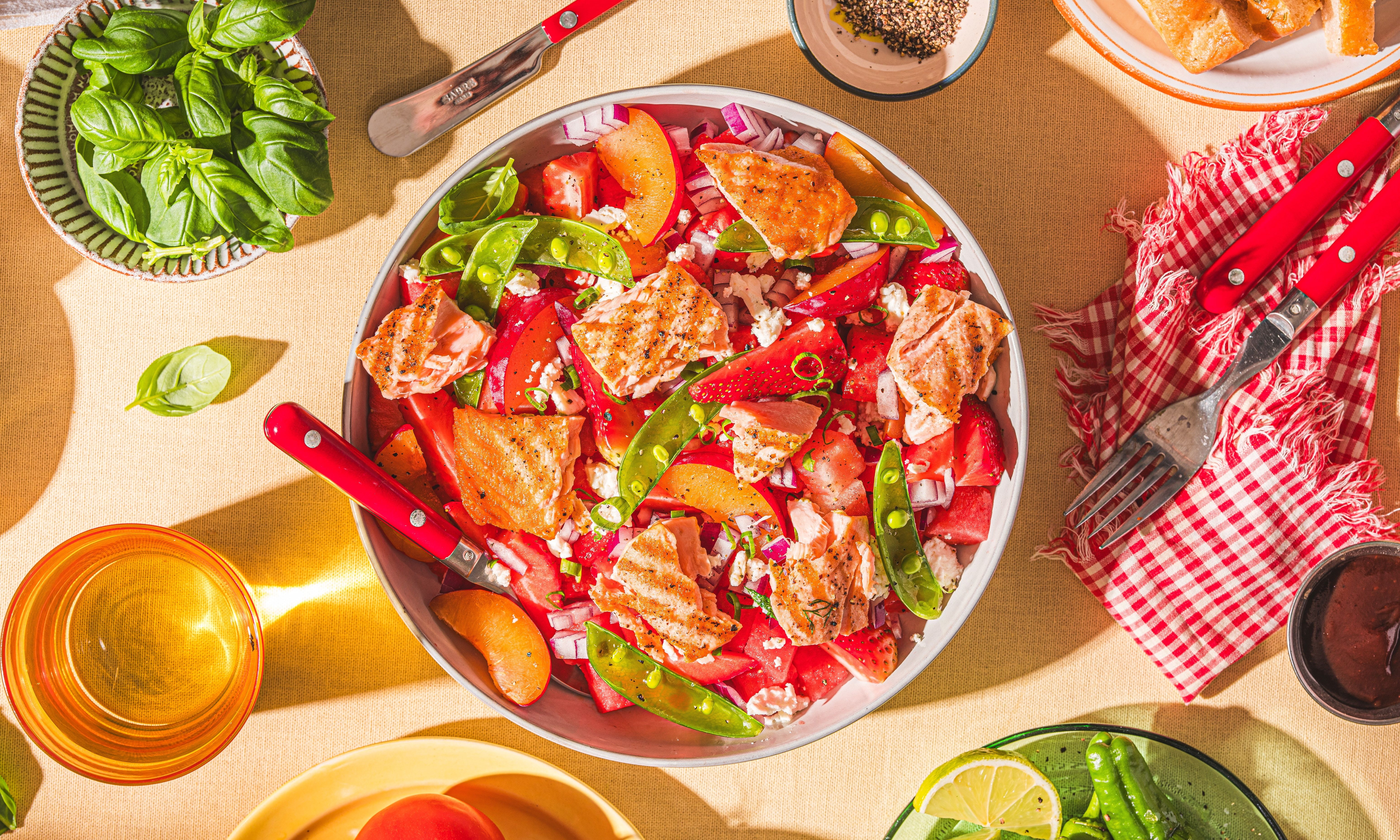 En salat med jordbær, vannmelon, og grønne blader i en hvit bolle, omgitt av ulike matretter og drikke på et bord.
