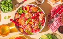 En salat med jordbær, vannmelon, og grønne blader i en hvit bolle, omgitt av ulike matretter og drikke på et bord.