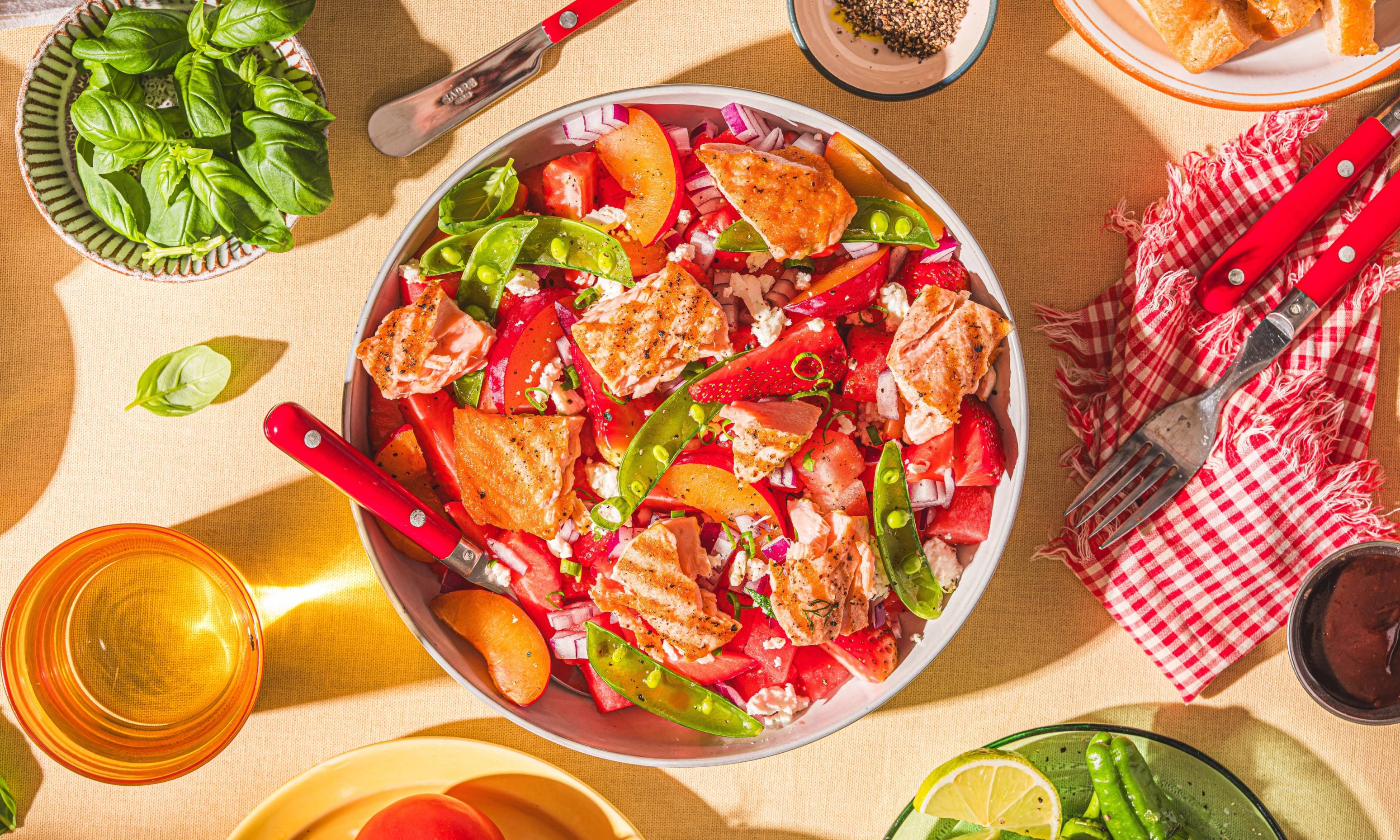 En salat med jordbær, vannmelon, og grønne blader i en hvit bolle, omgitt av ulike matretter og drikke på et bord.