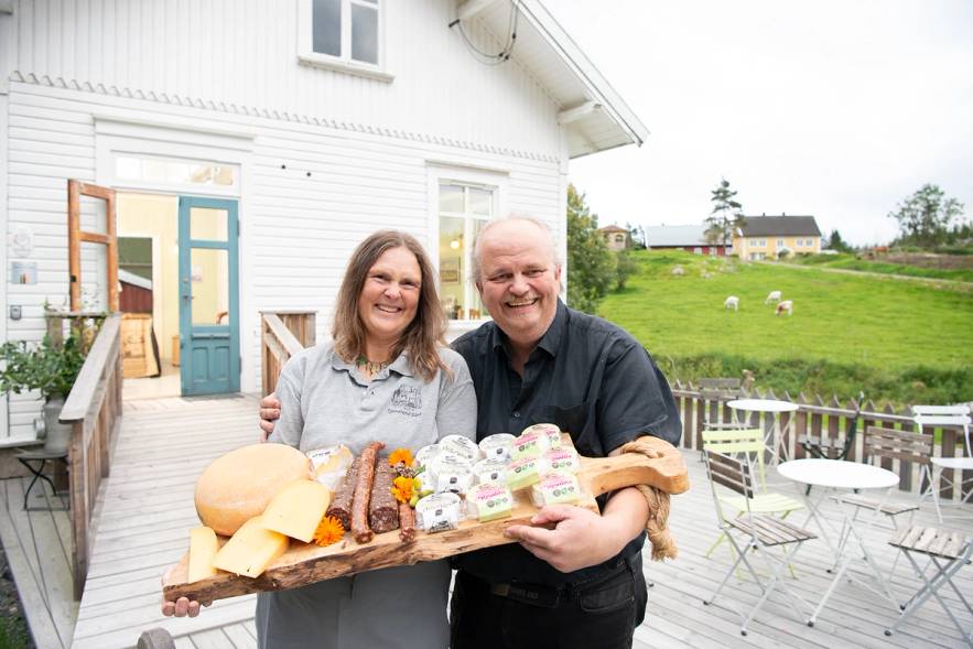 To personer holder en kake med lys og pynt foran en hvit husvegg på en veranda, med en grønn hage og noen sauer i bakgrunnen.