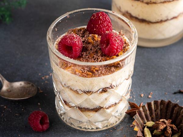 Et lagdelt dessert i et glass med krem, sjokoladekrønsj og friske bringebær på toppen.