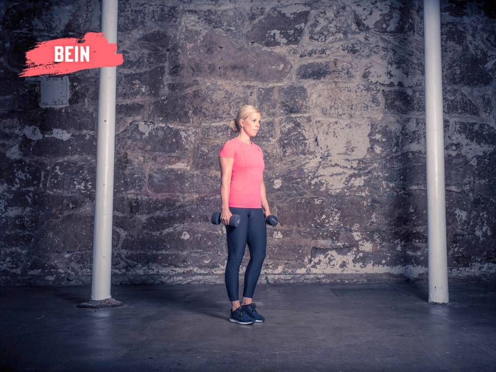 En kvinne står på ett bein mens hun holder en vekt i hver hånd foran kroppen, foran en murvegg.